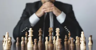 A person in a suit sitting behind a chessboard with their hands together in a "ready to play" position.
